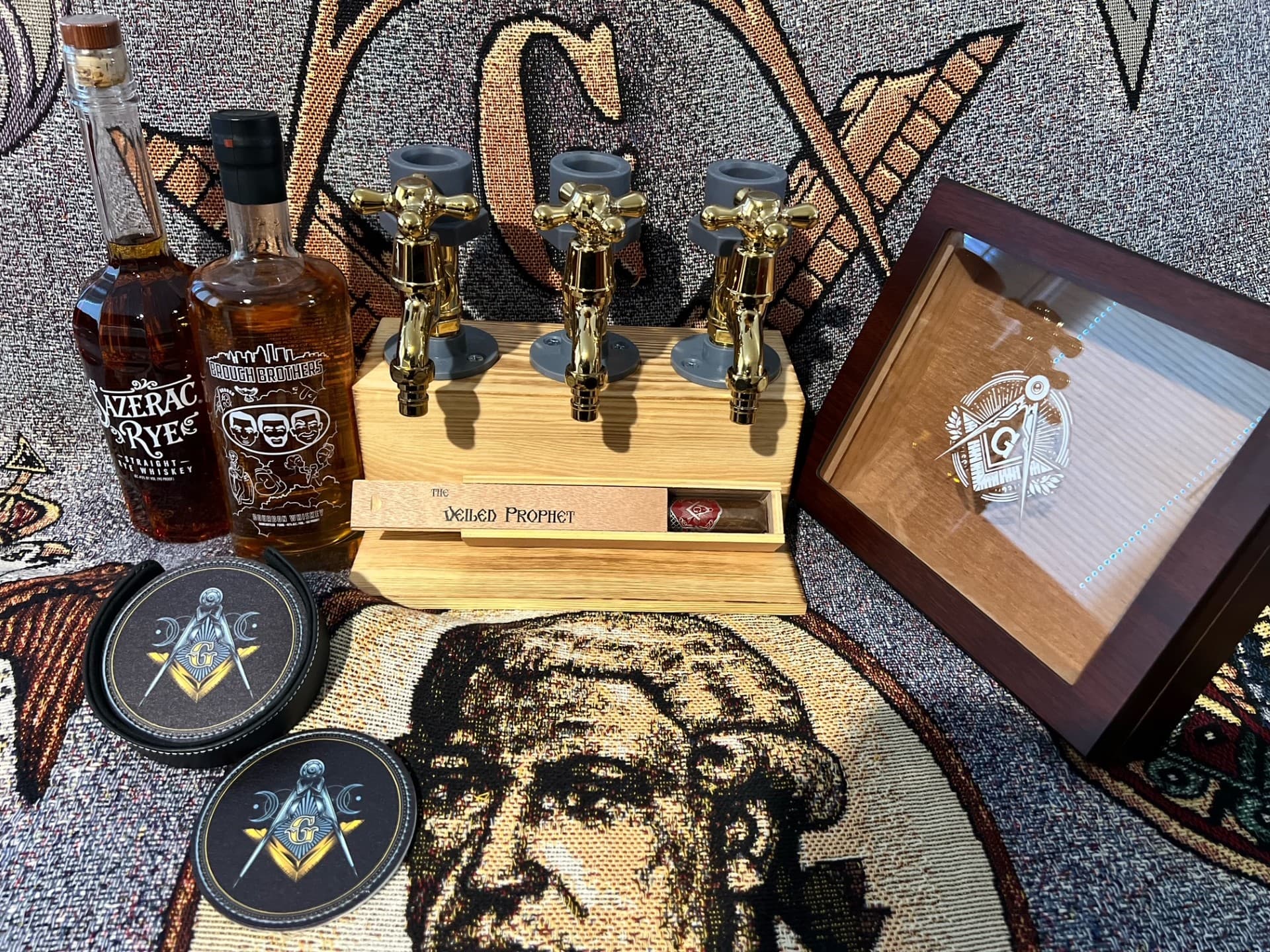 The Basket of Cheer — whiskey, gold-tap dispenser, cigars, and humidor arranged on a Masonic tapestry.